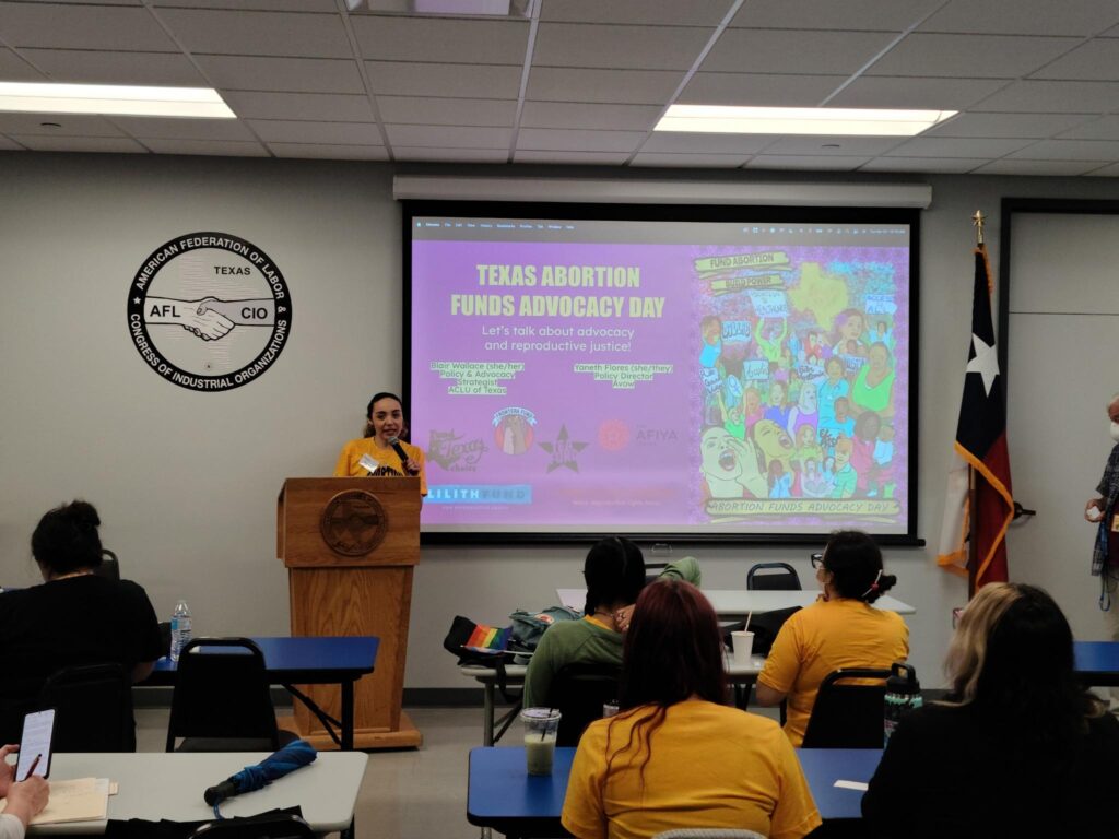 A person holding a microphone and wearing a yellow shirt that says “Abortion Forever” is standing at a podium in front of a slideshow that says, “Texas Abortion Funds Advocacy Day. Let’s talk about advocacy and reproductive justice!”