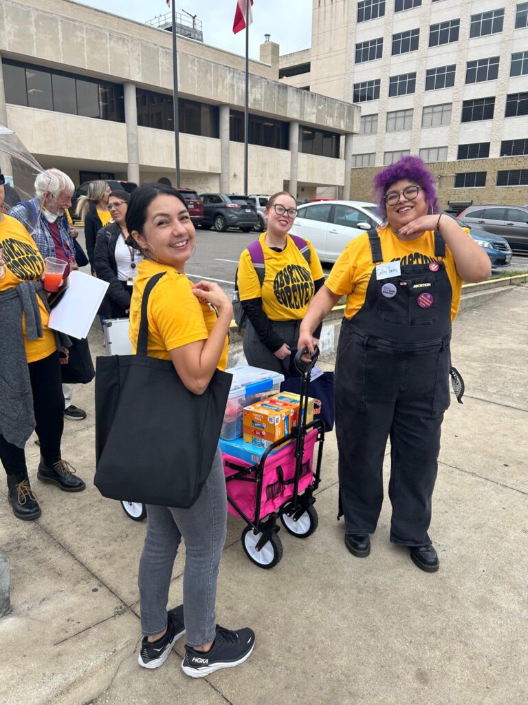 3 people wearing yellow “Abortion Forever” shirts are smiling on a sidewalk with a wagon of supplies.